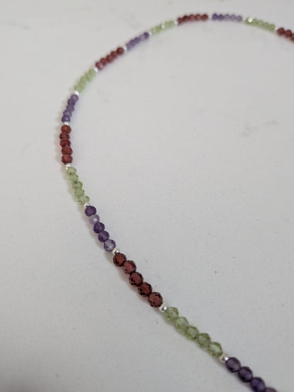A beaded necklace made of small faceted gemstone beads in a rainbow sequence of colors, including purple, red, green, and blue, arranged in a curved shape on a plain light background.