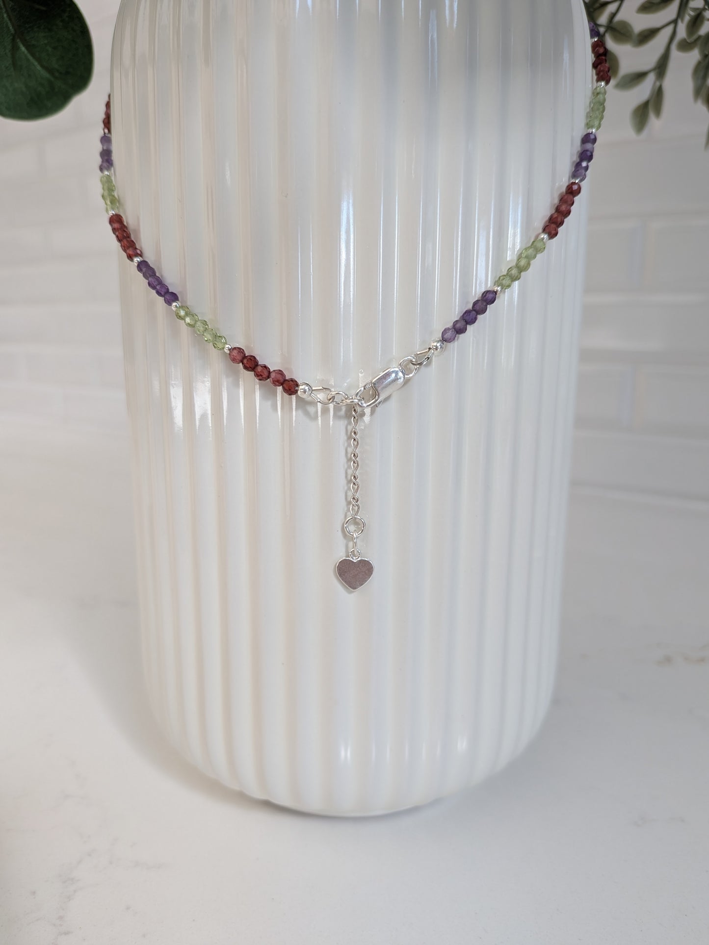 A beaded chakra necklace with a silver clasp and heart charm, displayed on a white ribbed vase.