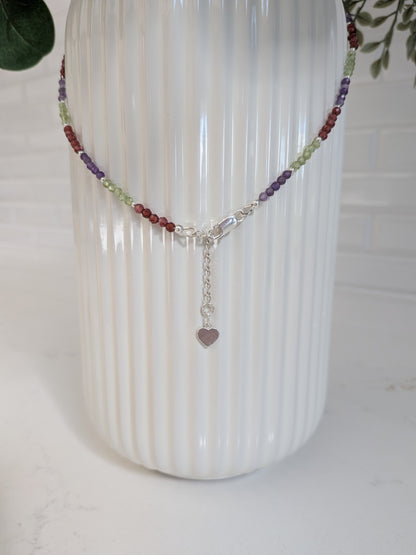 A beaded chakra necklace with a silver clasp and heart charm, displayed on a white ribbed vase.