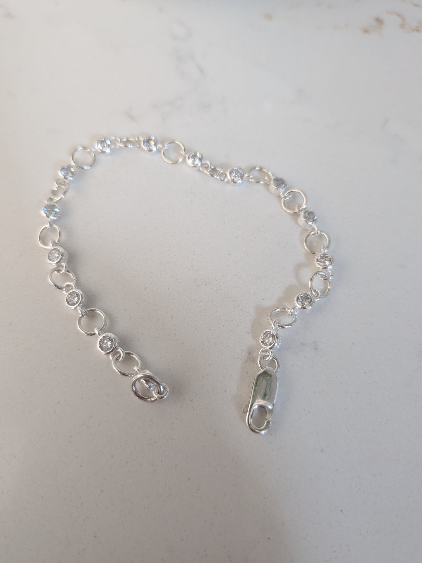 A delicate silver chain bracelet with small clear faceted gemstones and circular links, featuring a lobster clasp closure, displayed on a light gray surface.