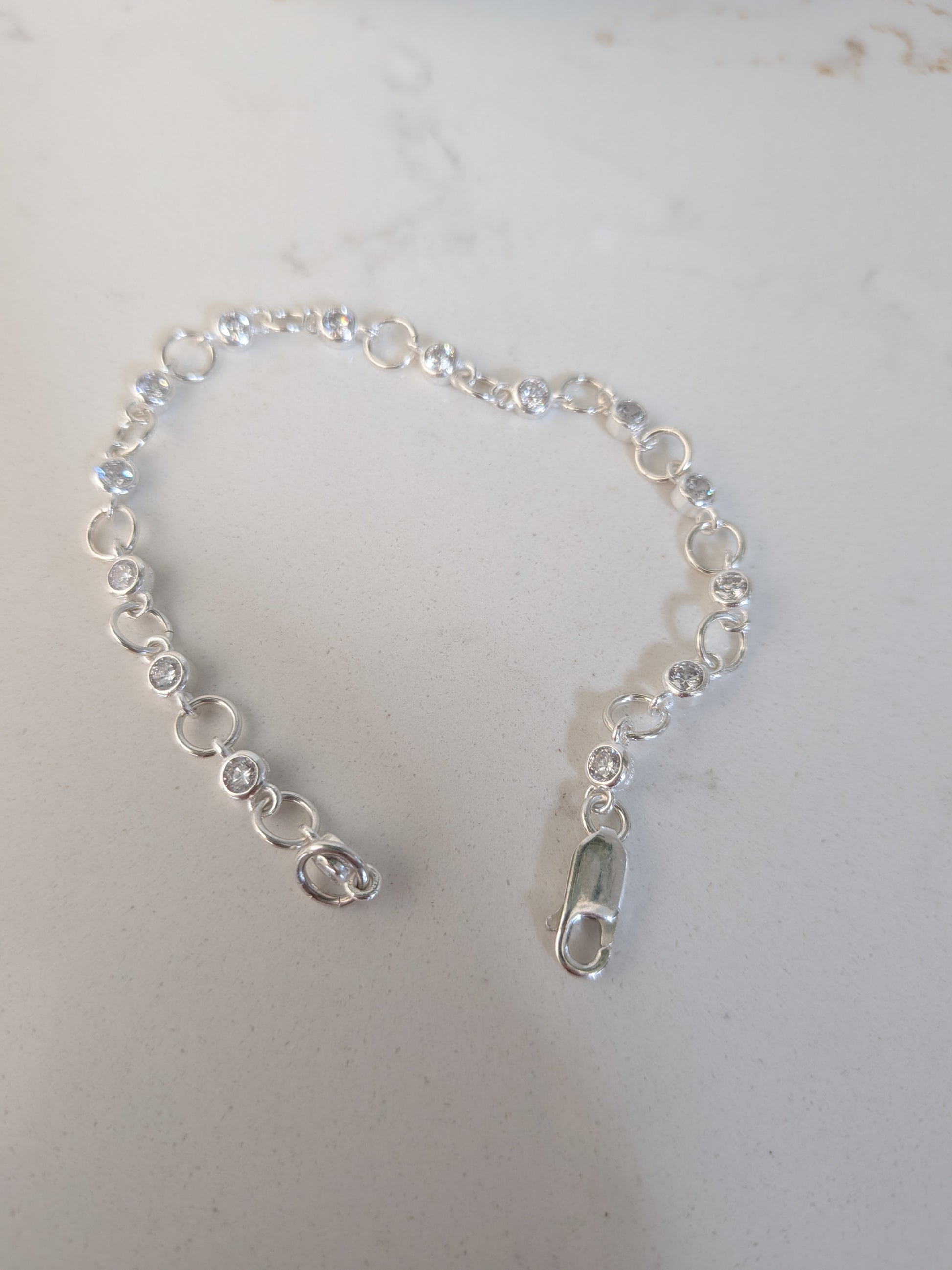 A delicate silver chain bracelet with small clear faceted gemstones and circular links, featuring a lobster clasp closure, displayed on a light gray surface.