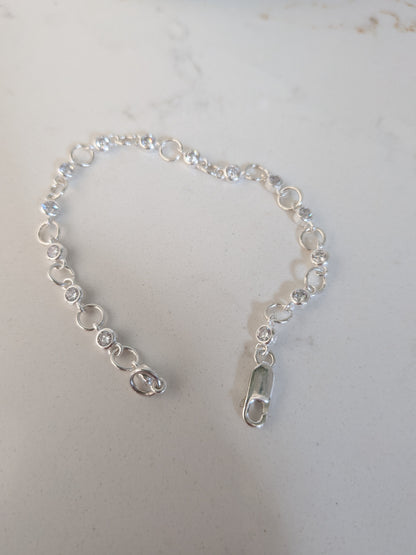 A delicate silver chain bracelet with small clear faceted gemstones and circular links, featuring a lobster clasp closure, displayed on a light gray surface.