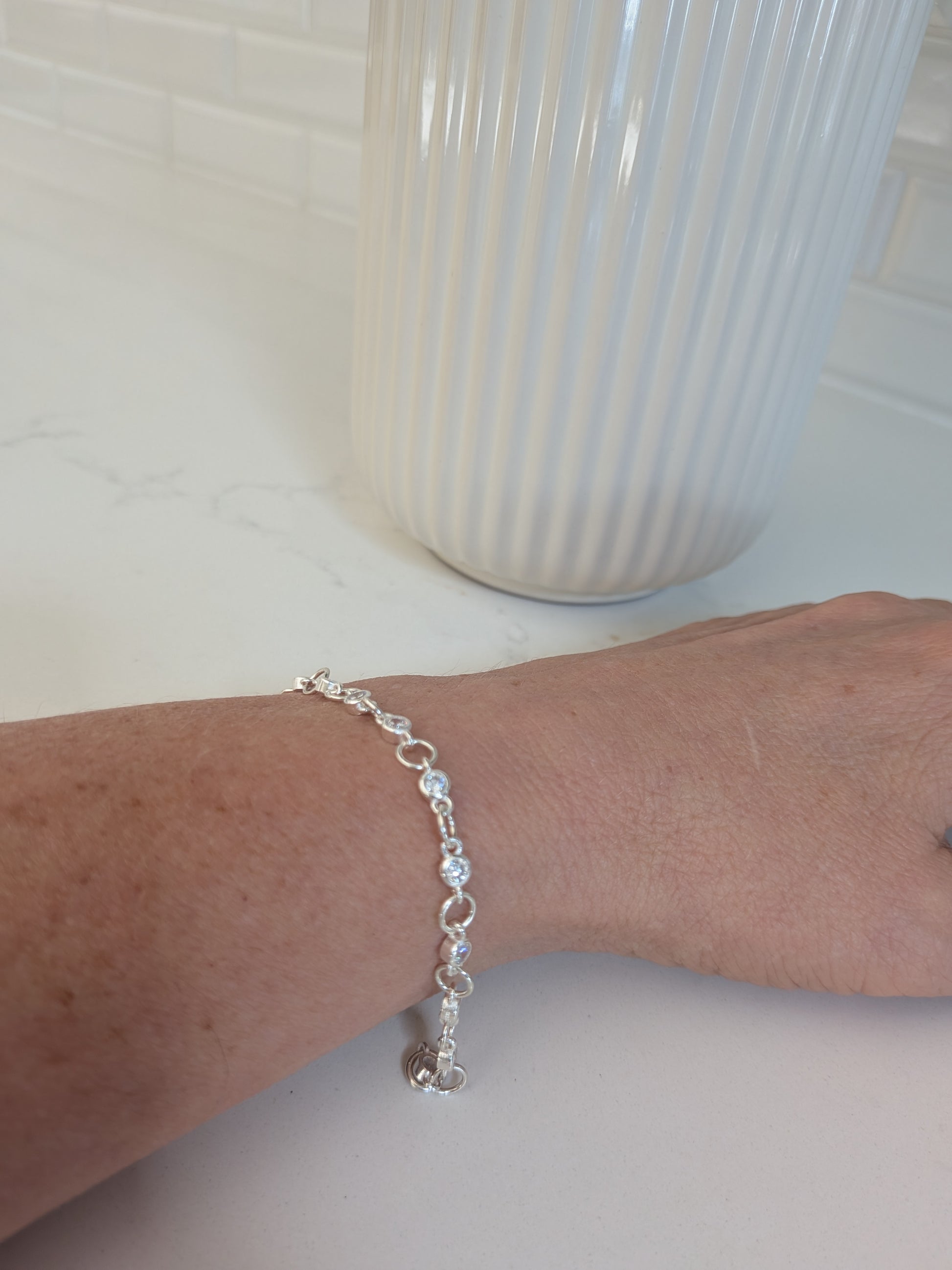 A delicate silver chain bracelet with clear faceted beads worn on a person's wrist, shown against a white countertop with a ribbed white vase in the background.
