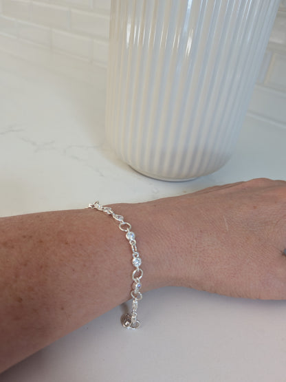 A delicate silver chain bracelet with clear faceted beads worn on a person's wrist, shown against a white countertop with a ribbed white vase in the background.