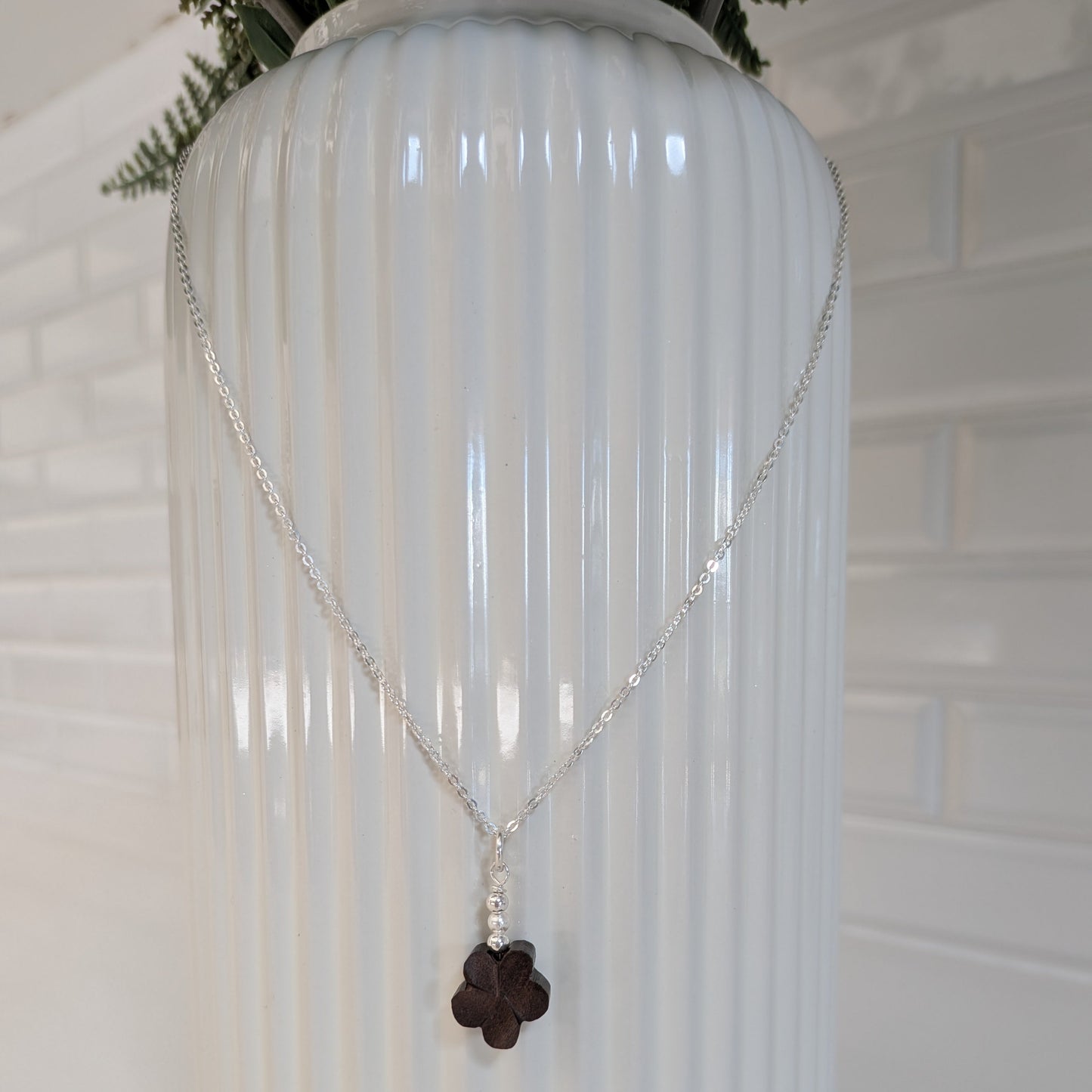 A silver chain necklace with a dark brown wood flower pendant and Swarovski pearls, draped over a white ribbed ceramic vase against a white brick wall background.