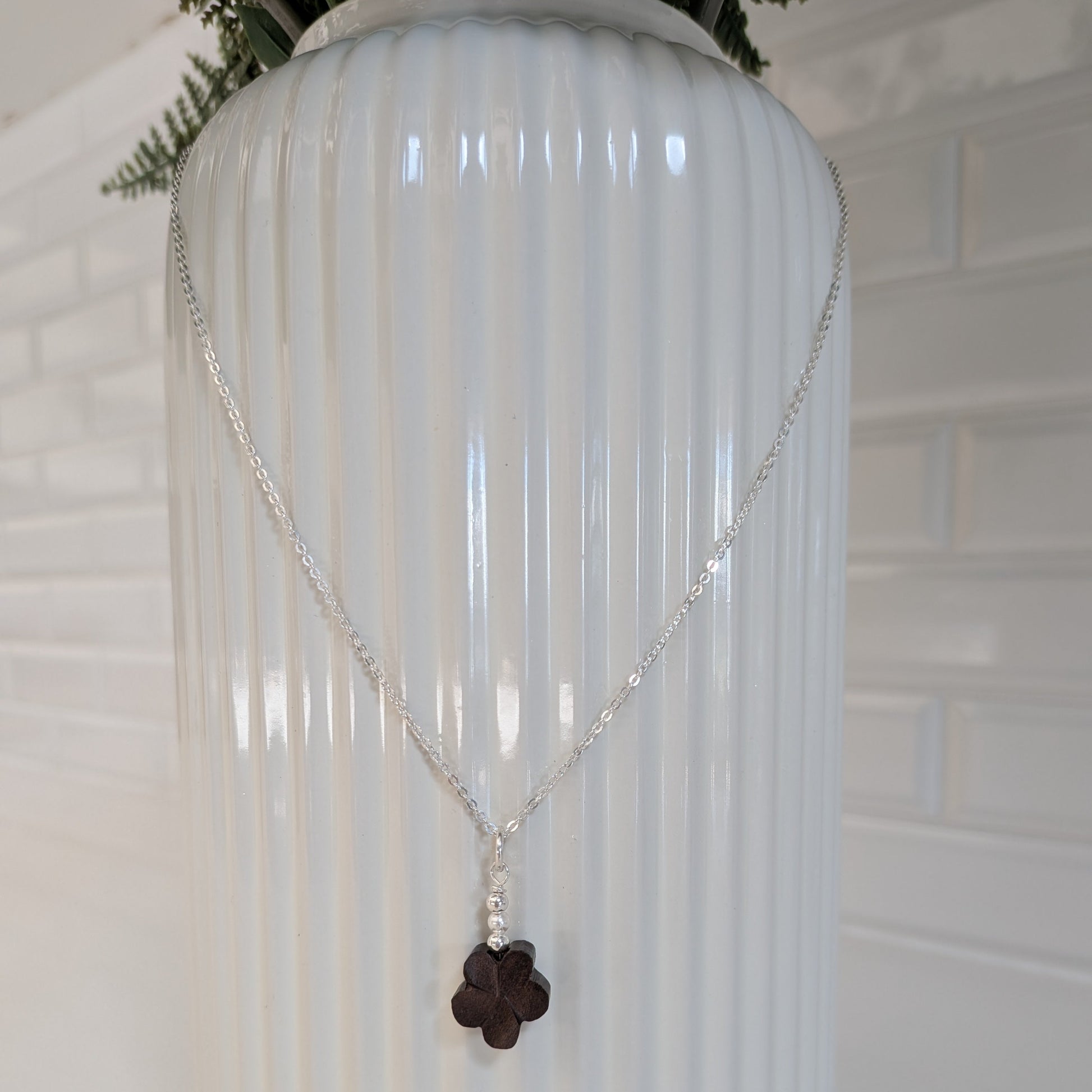 A silver chain necklace with a dark brown wood flower pendant and Swarovski pearls, draped over a white ribbed ceramic vase against a white brick wall background.