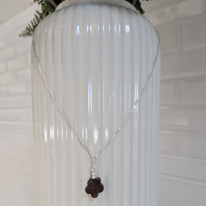 A silver chain necklace with a dark brown wood flower pendant and Swarovski pearls, draped over a white ribbed ceramic vase against a white brick wall background.