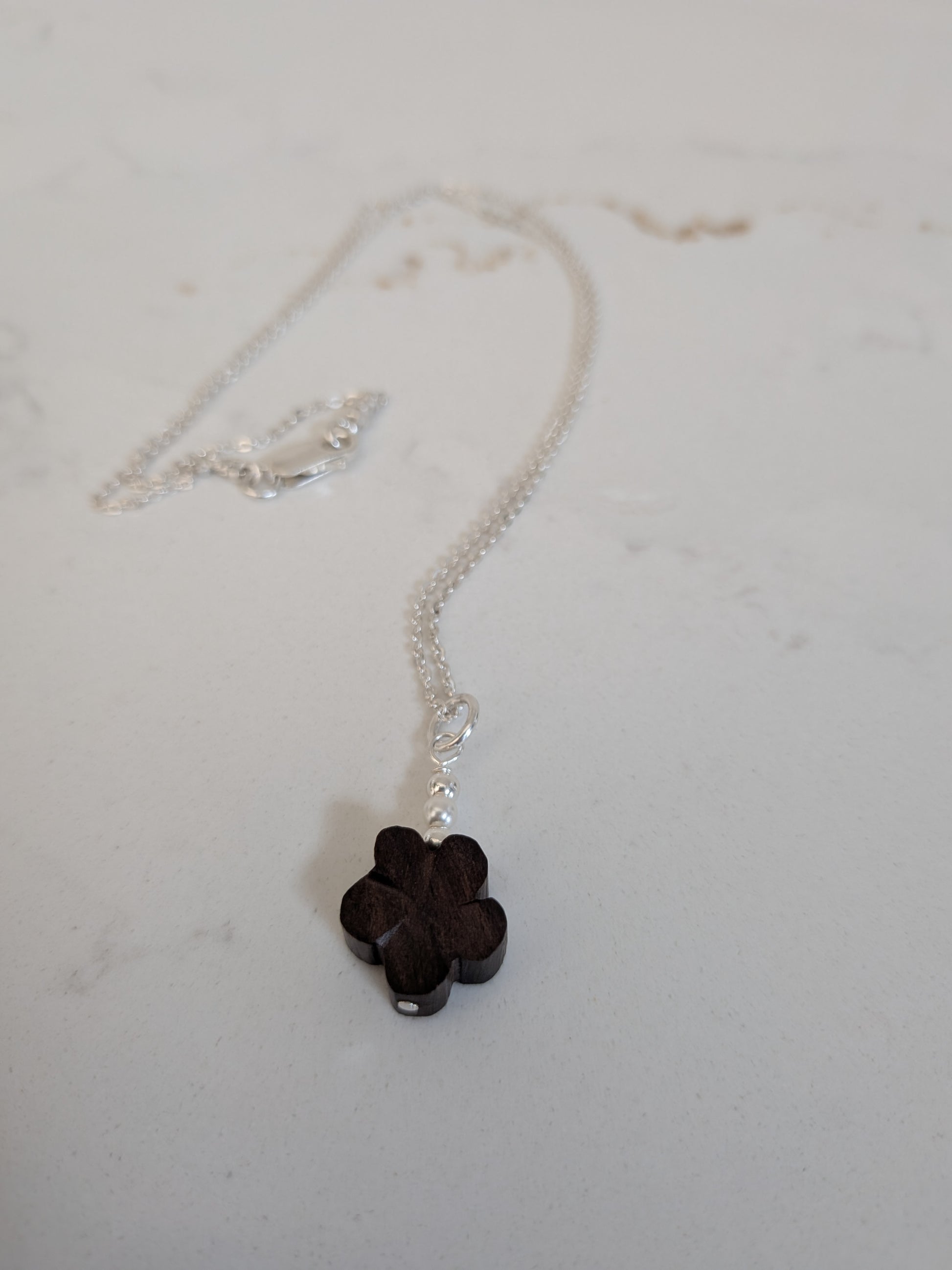 A silver chain necklace with a small dark brown wooden flower and Swarovski pearls pendant, resting on a light gray surface.