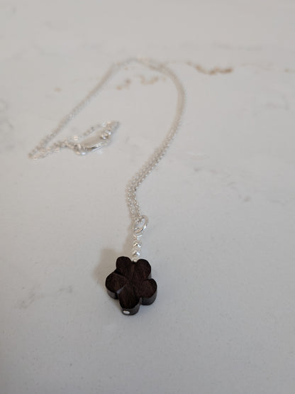 A silver chain necklace with a small dark brown wooden flower and Swarovski pearls pendant, resting on a light gray surface.