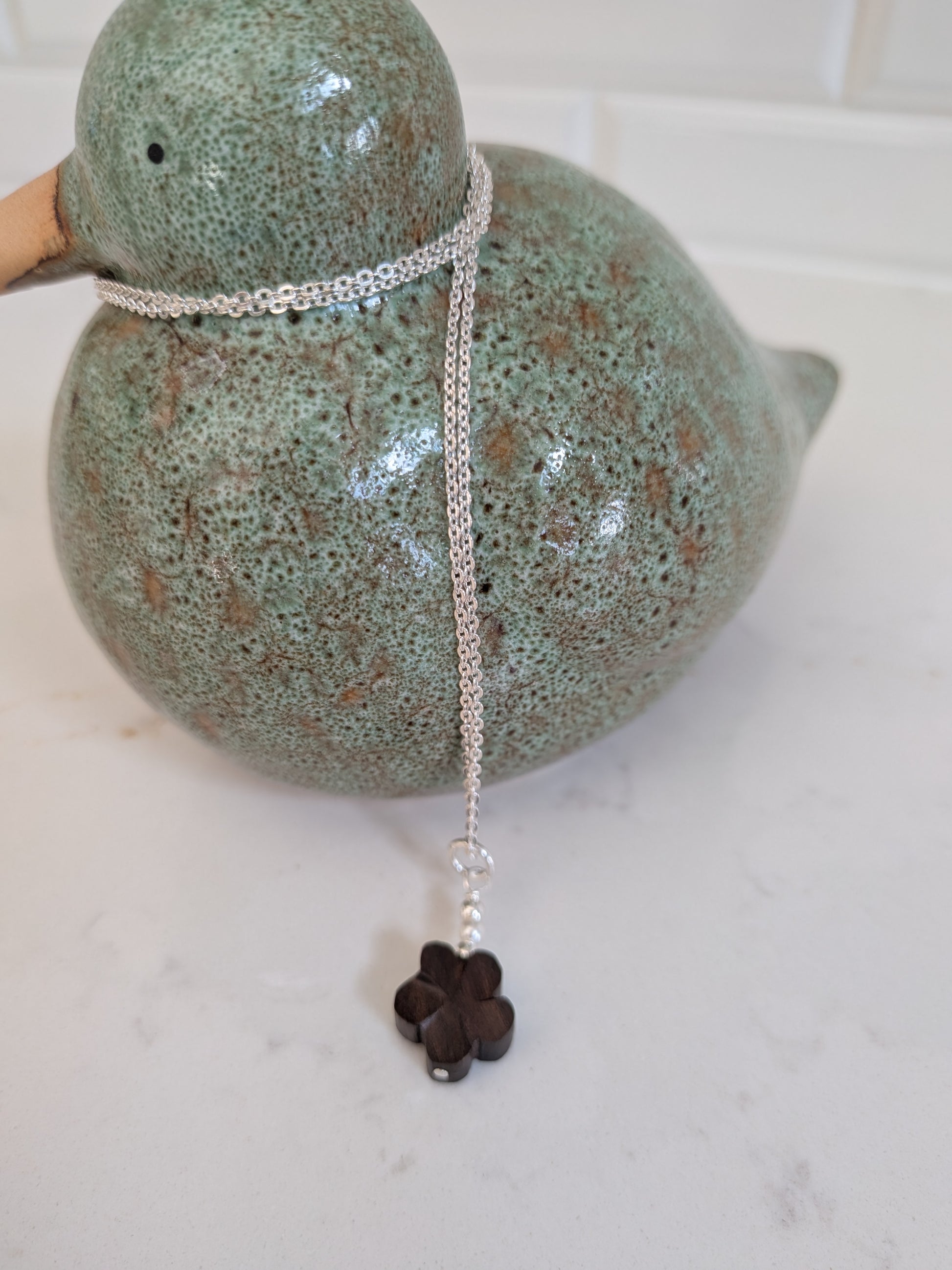 A silver chain necklace with a dark brown wooden flower and Swarovski pearls pendant, draped over a speckled green ceramic duck figurine on a white marble surface.