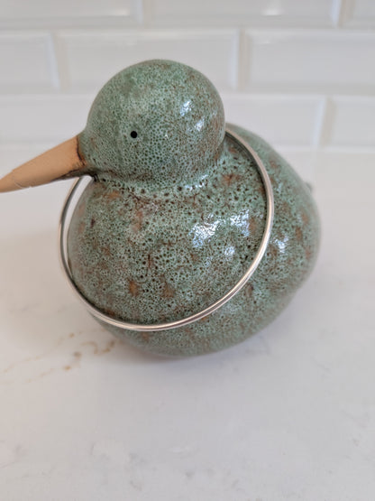 A ceramic duck-shaped container with a speckled sage green and brown glaze, featuring a light wood beak and a silver metal ring handle, set on a white marble countertop with a white subway tile backsplash.
