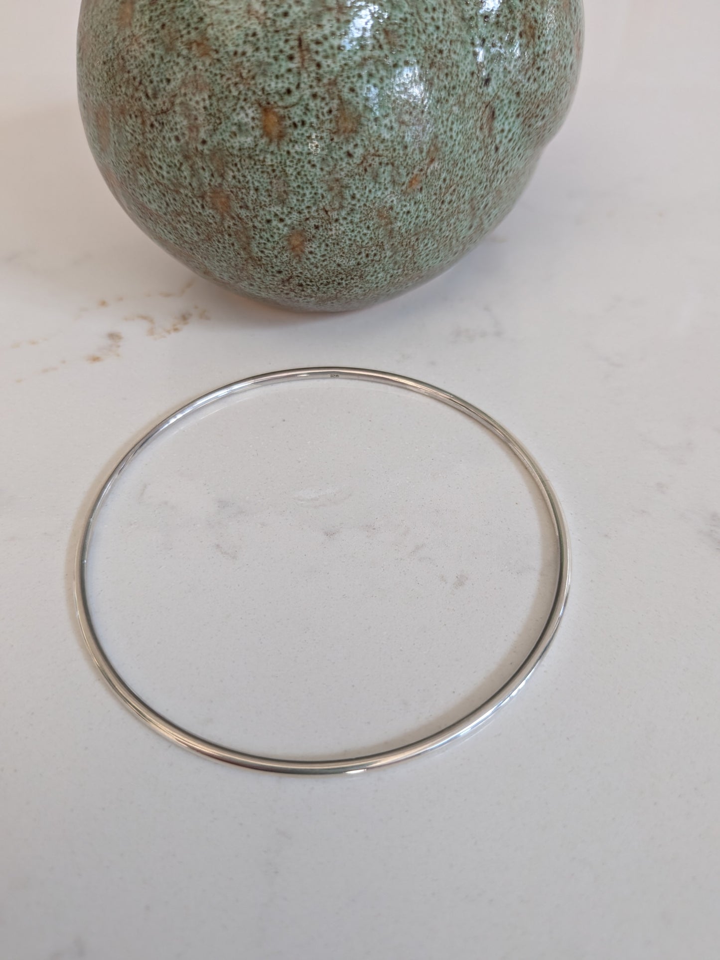 A thin silver-toned metal bangle bracelet displayed on a light off-white surface, with a green speckled ceramic vase partially visible in the background.