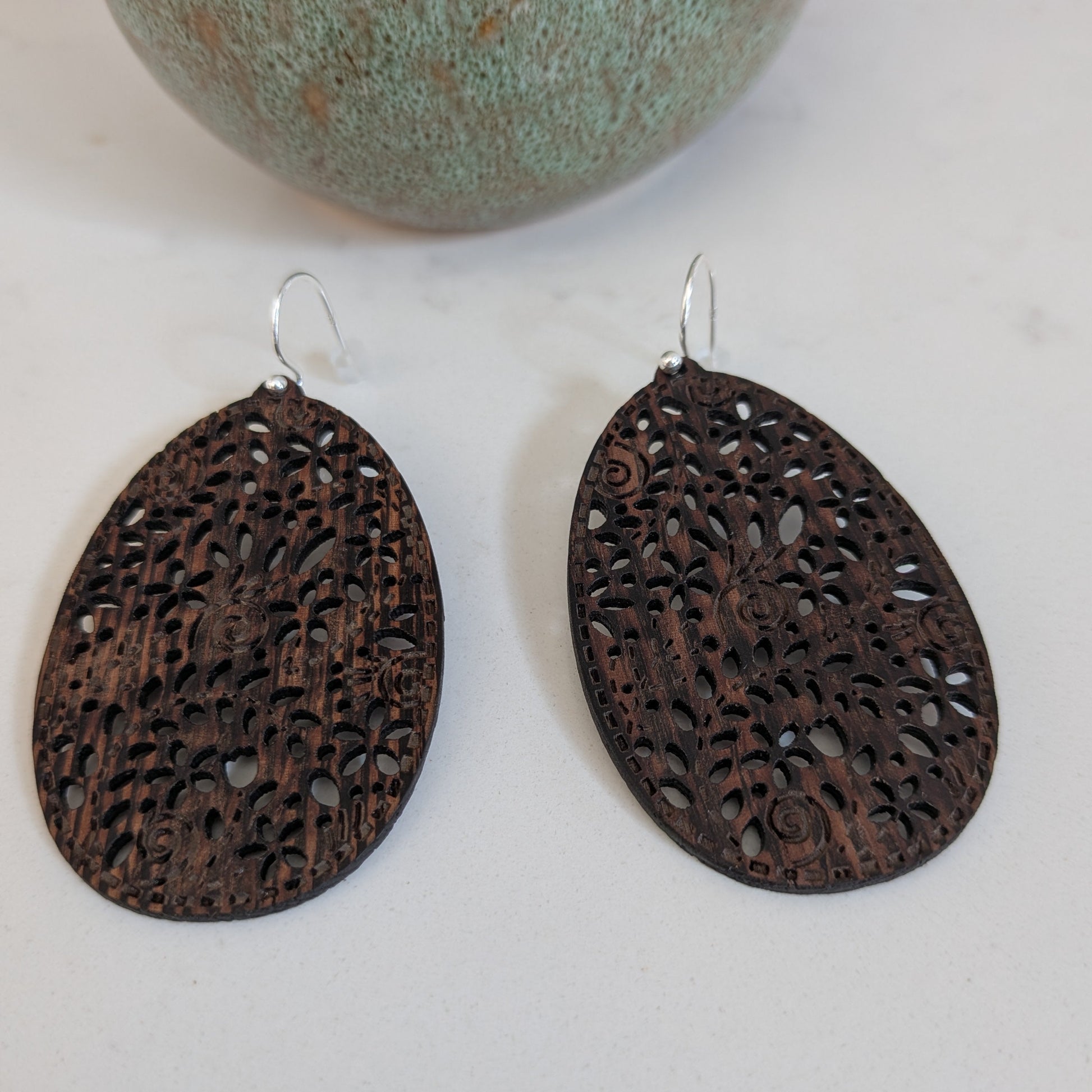 A pair of dark brown teardrop-shaped wooden earrings with intricate laser-cut floral patterns and silver-tone hooks, displayed on a light surface with a decorative green ceramic vase in the background.