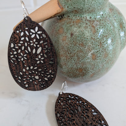 A pair of dark brown laser-cut wooden dangle earrings with silver fishhook wires, displayed on a white marble surface next to a green ceramic vase.