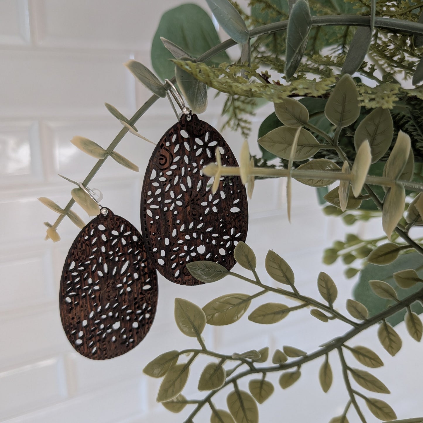 Two dark brown laser-cut metal teardrop earrings with intricate white cutout patterns, hanging from a decorative branch with green leaves against a white paneled wall.