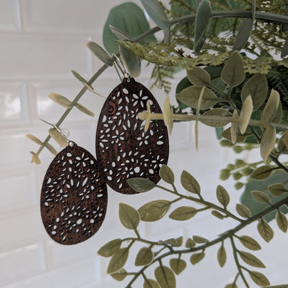 Two dark brown laser-cut metal teardrop earrings with intricate white cutout patterns, hanging from a decorative branch with green leaves against a white paneled wall.