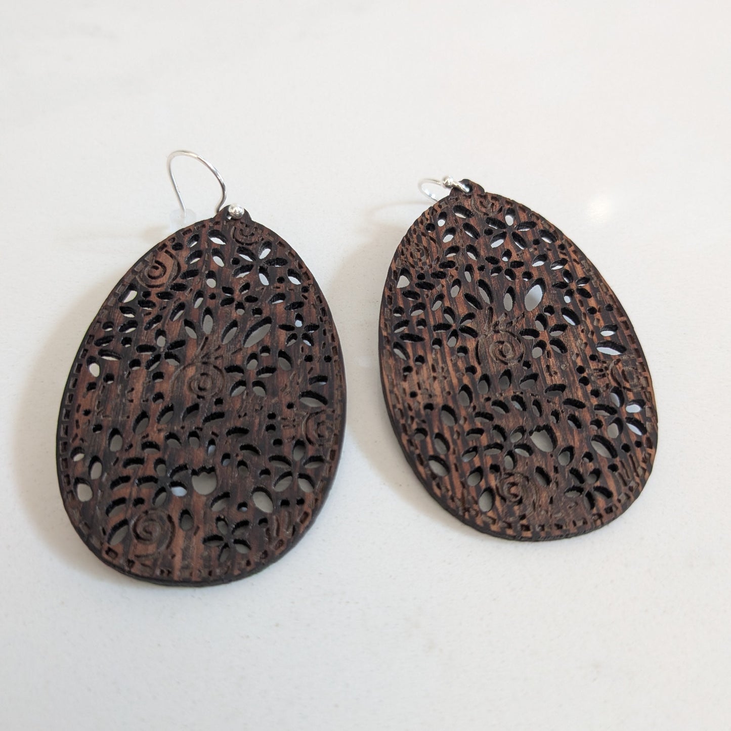 A pair of dark brown teardrop-shaped wooden earrings with intricate laser-cut filigree patterns, displayed on a light off-white background.