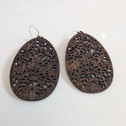 A pair of dark brown teardrop-shaped wooden earrings with intricate laser-cut filigree patterns, displayed on a light off-white background.