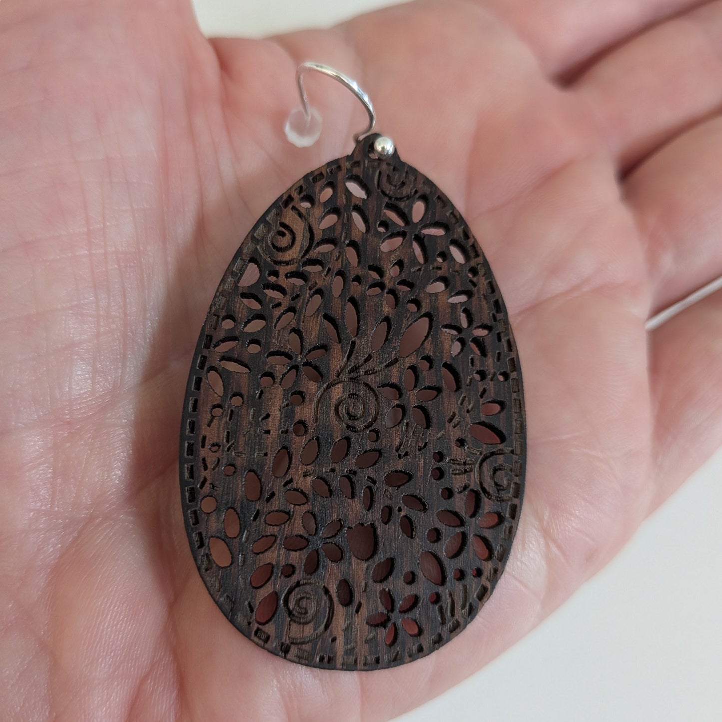 A dark brown teardrop-shaped wooden pendant with intricate floral filigree cutouts, held in a person's hand. The pendant features a small silver metal loop at the top.