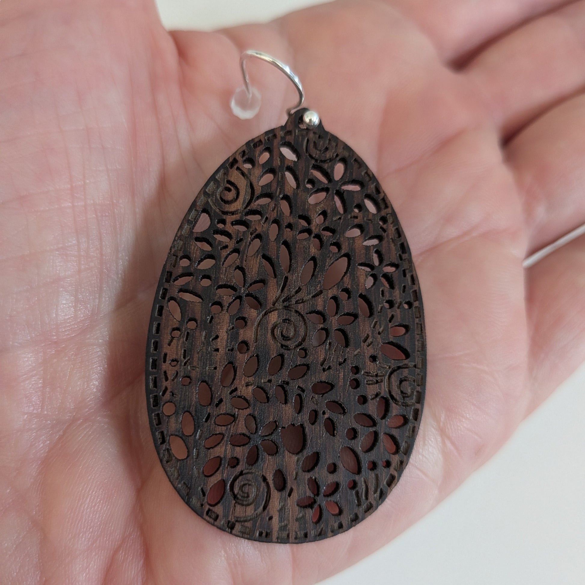 A dark brown teardrop-shaped wooden pendant with intricate floral filigree cutouts, held in a person's hand. The pendant features a small silver metal loop at the top.