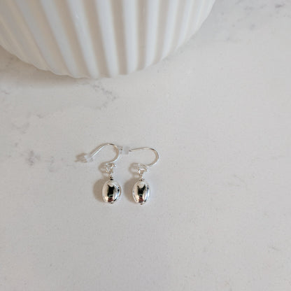 A pair of silver puffed oval drop earrings displayed on a light marble surface next to a white ribbed ceramic container.