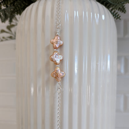 A silver rolo chain bracelet with three vertical champagne four leaf clover-pearls, hanging against a white ribbed ceramic vase.