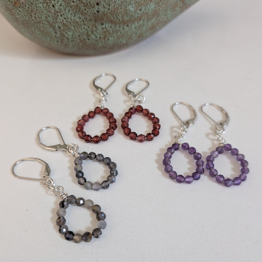 Three pairs of beaded hoop earrings with silver fishhook backings are displayed on a light surface. The earrings feature faceted beads in dark gray, deep burgundy red, and purple.