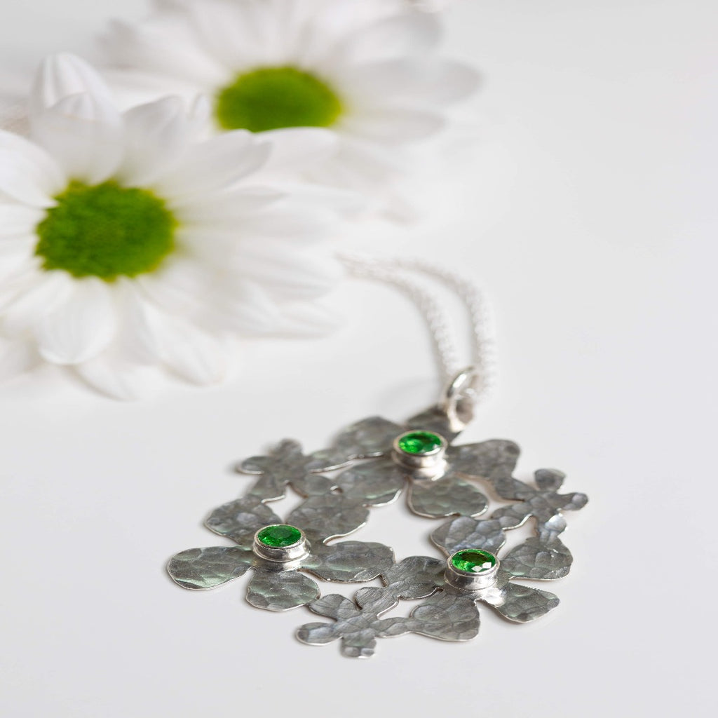 Image of a Sterling Silver Handmade Daisy Delight Necklace adorned with vibrant tsavorite garnets. This stunning piece features a 20-gauge sterling silver pendant, tube-set tsavorite garnets, and a sterling silver wheat chain with a lobster clasp. Inspired by nature, the delicate daisy cluster design and captivating gemstones evoke a sense of beauty and transformation, perfect for reconnecting with nature's allure and awakening the beauty within.