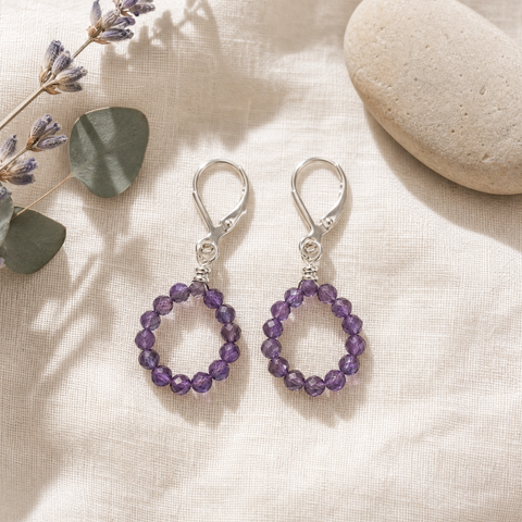 A pair of teardrop-shaped drop earrings made of purple beads with silver hooks, displayed on a textured beige fabric alongside dried lavender and a smooth stone.