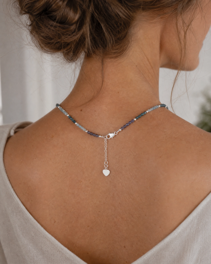 A close-up back view of a woman wearing a delicate beaded necklace with a small silver heart dangle, styled with a light beige top.