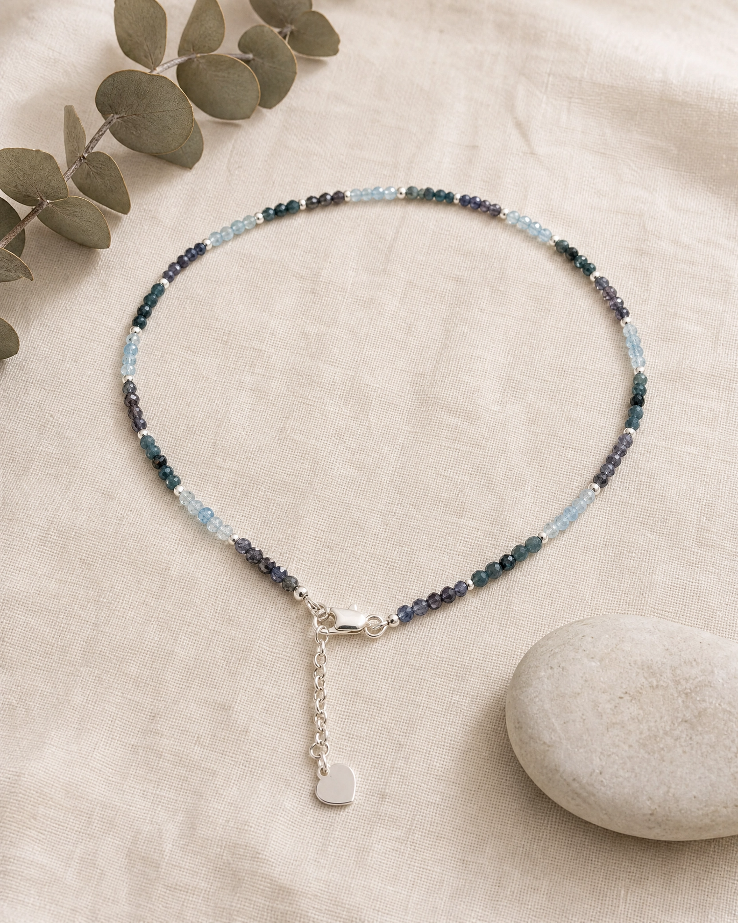 A delicate beaded necklace with small blue, green, and purple beads, featuring a silver lobster clasp and a dangling silver heart charm, displayed on a beige linen fabric with eucalyptus leaves and a stone.