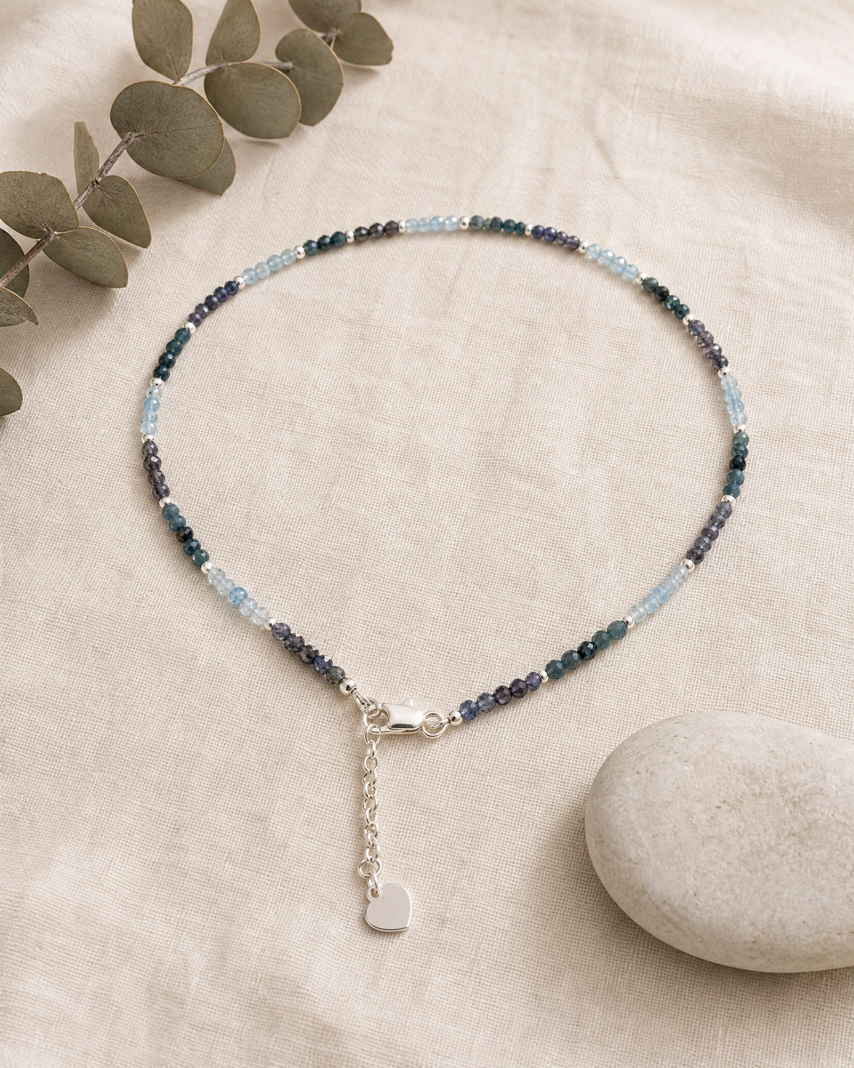 A delicate beaded necklace with small blue, green, and purple beads, featuring a silver lobster clasp and a dangling silver heart charm, displayed on a beige linen fabric with eucalyptus leaves and a stone.