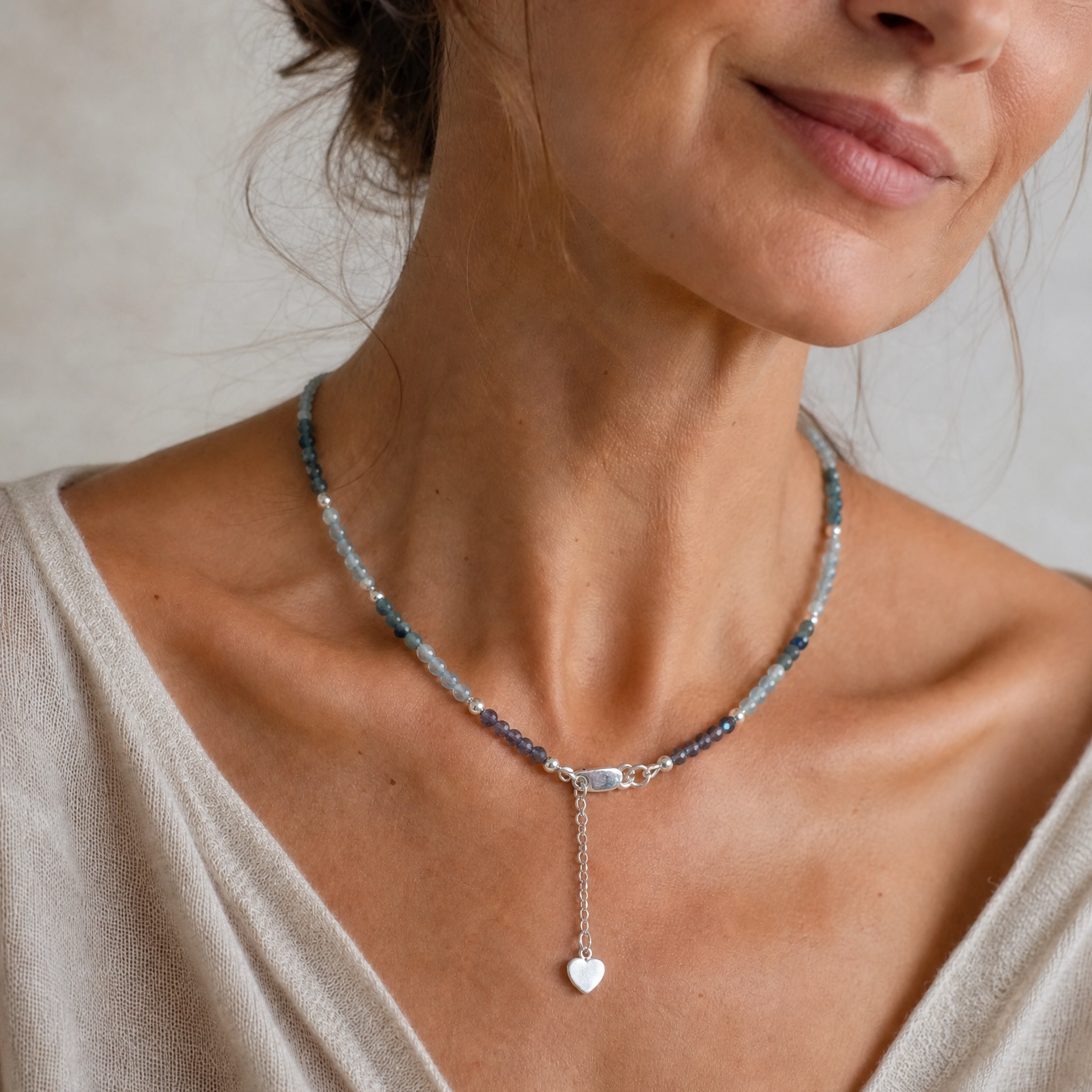 A close-up of a woman wearing a delicate beaded necklace with a silver-toned heart pendant, paired with a light beige V-neck top.
