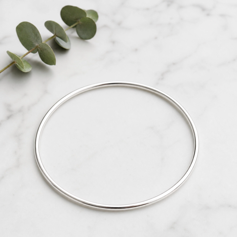 A thin, polished silver bangle bracelet displayed on a white marble surface with a green eucalyptus sprig in the top-left corner.