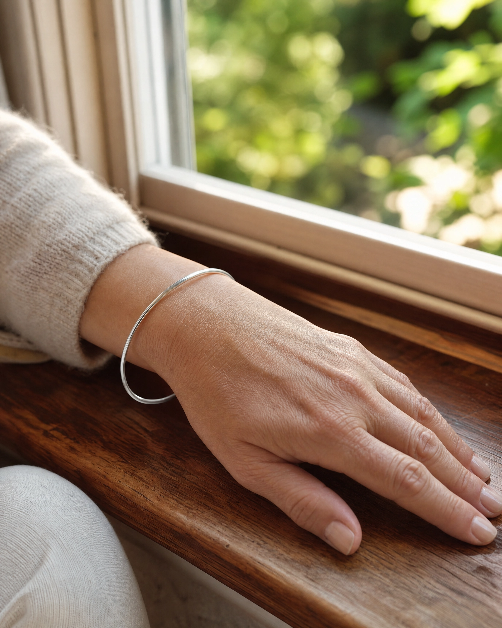 A thin silver bangle bracelet worn on a person's wrist, resting on a dark wooden windowsill with a window and green foliage visible in the background.