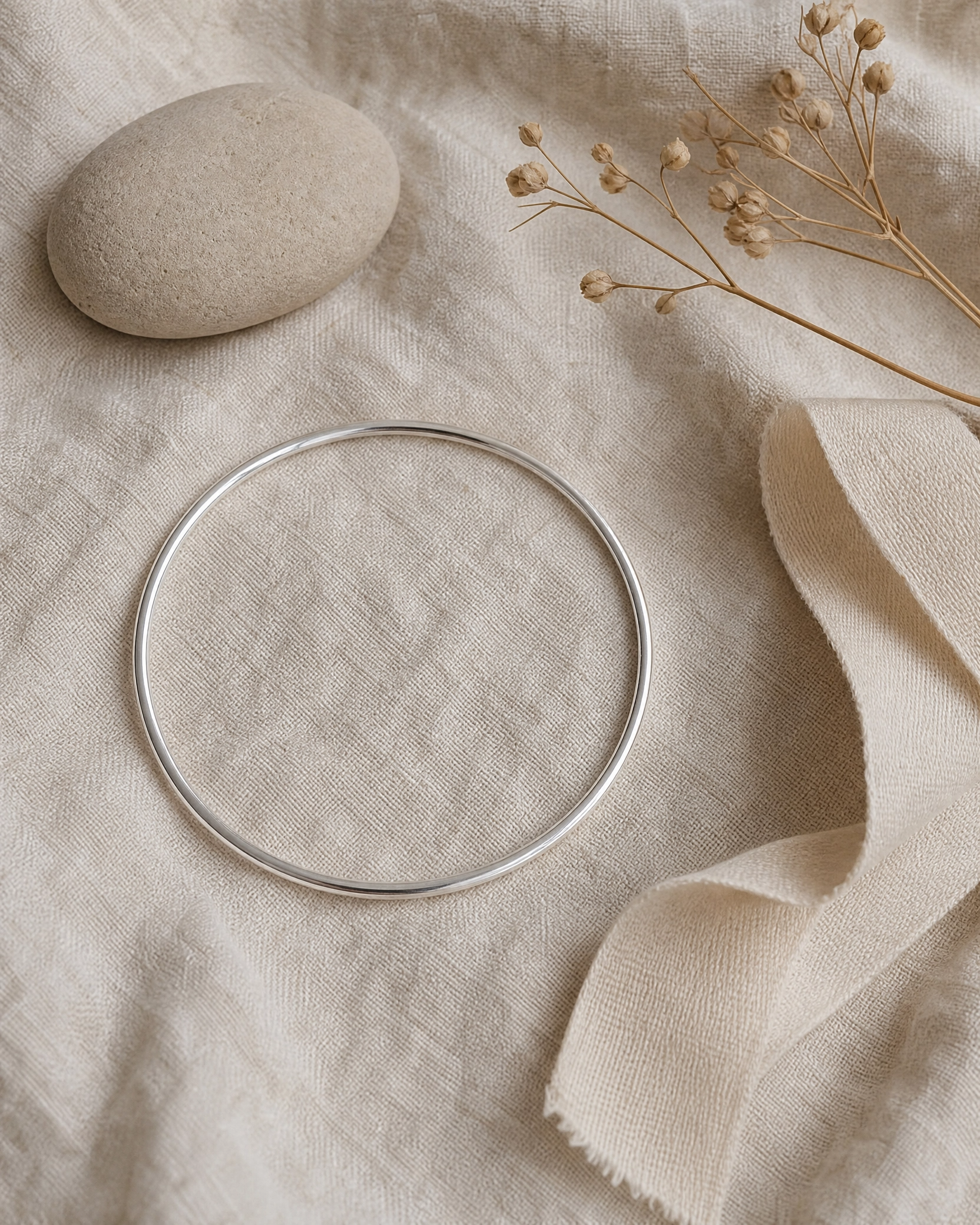 A thin silver-toned metal bangle bracelet is arranged on a textured beige linen fabric with a smooth stone, dried flowers, and a folded ribbon.