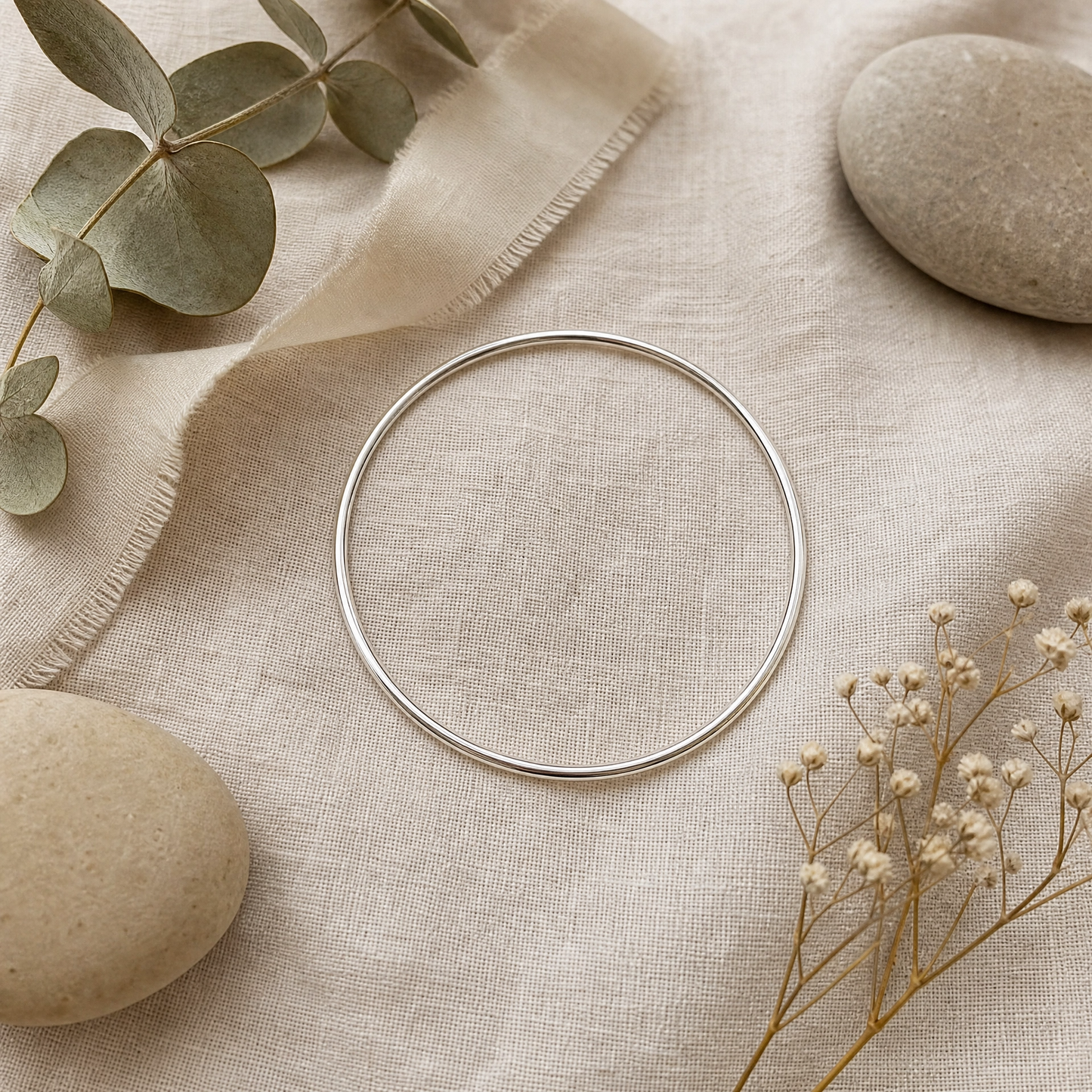 A thin silver bangle bracelet is displayed on a textured beige linen cloth, styled with natural stones, dried baby's breath, and eucalyptus leaves.