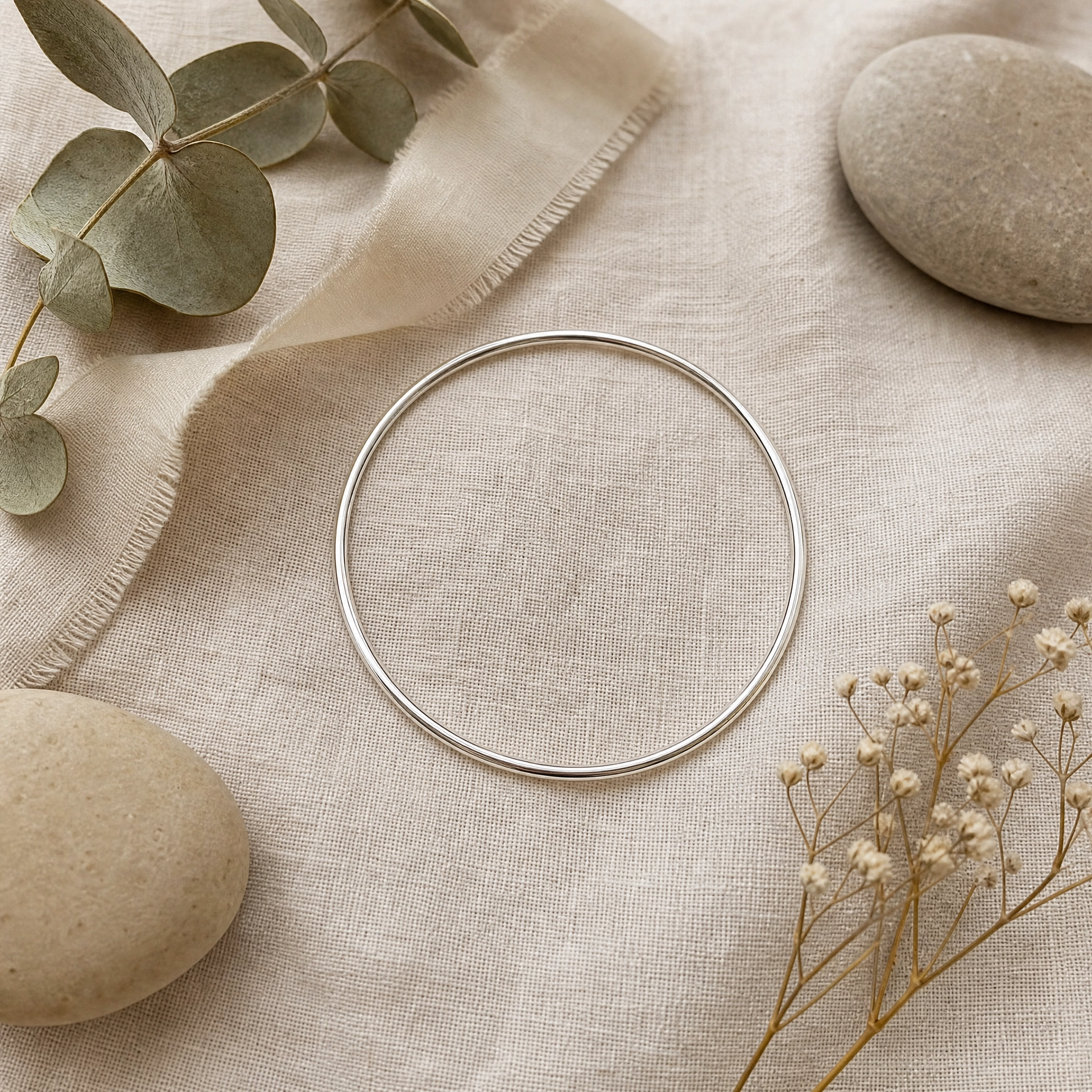 A thin silver bangle bracelet is displayed on a textured beige linen cloth, styled with natural stones, dried baby's breath, and eucalyptus leaves.