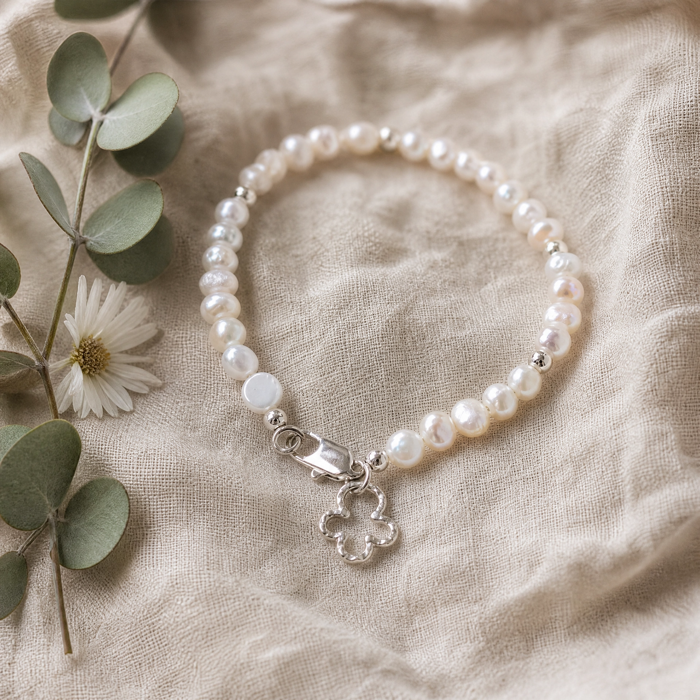 A beaded bracelet made of white freshwater pearls and silver beads, featuring a silver clasp and a dangling silver flower charm, styled on a beige linen fabric with eucalyptus leaves and a small white flower.