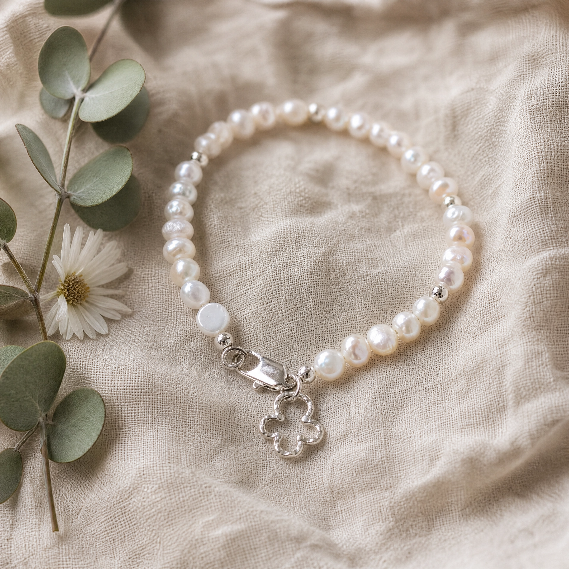 A beaded bracelet made of white freshwater pearls and silver beads, featuring a silver clasp and a dangling silver flower charm, styled on a beige linen fabric with eucalyptus leaves and a small white flower.
