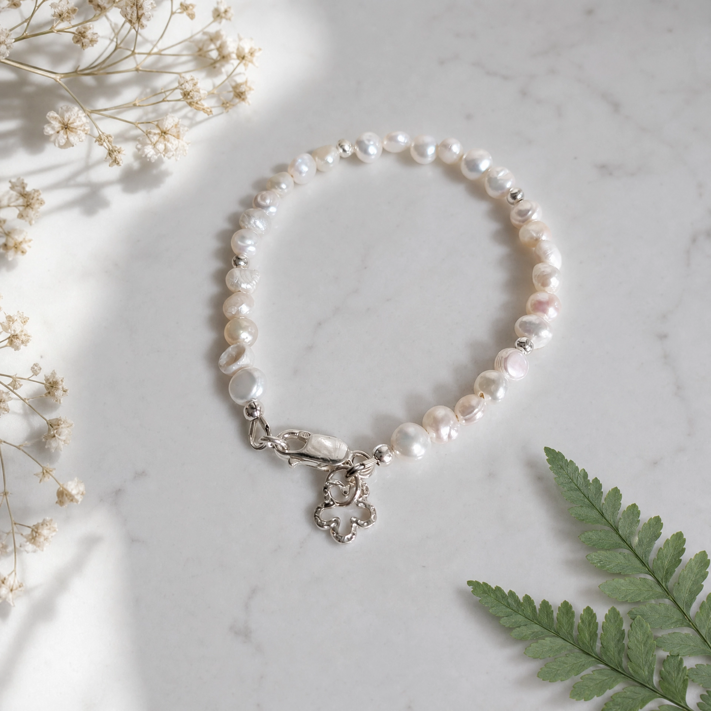 A beaded bracelet made of irregular white button pearls with a silver clasp and flower charm, styled on a white marble surface with dried flowers and a green fern leaf.