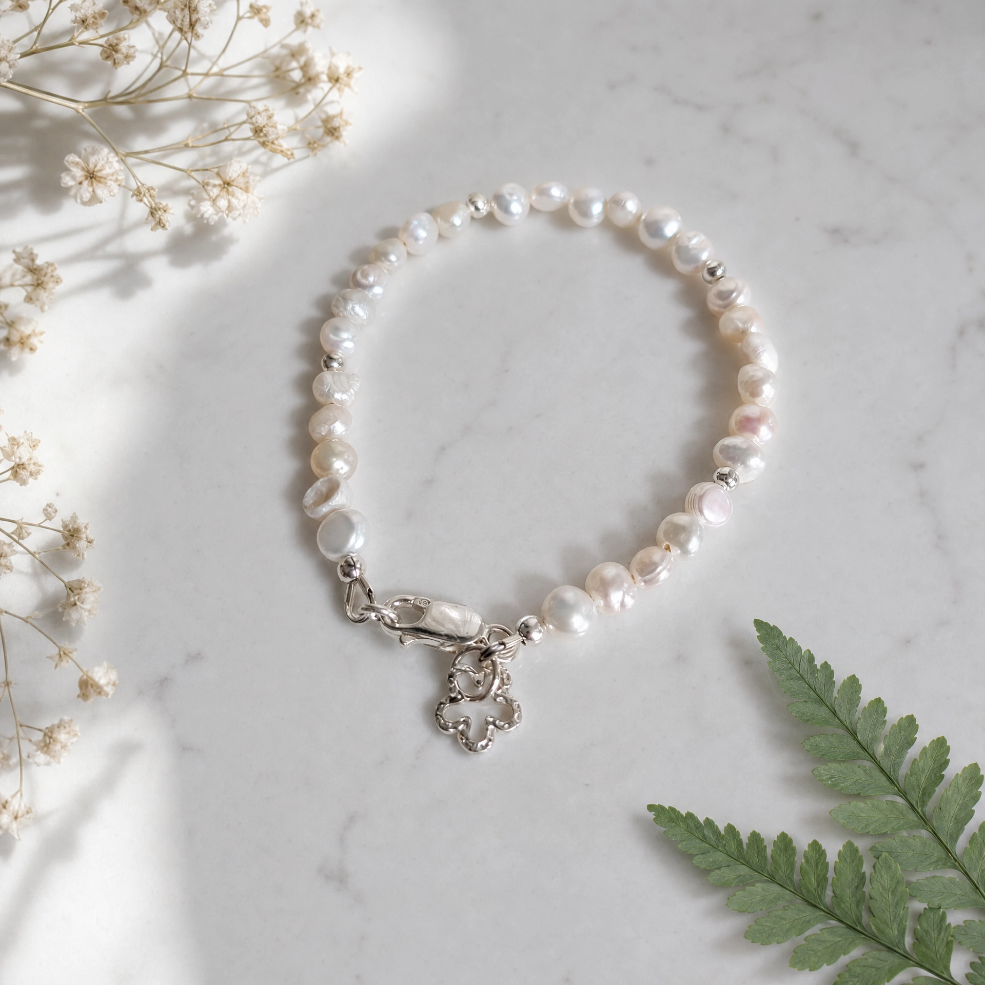 A beaded bracelet made of irregular white button pearls with a silver clasp and flower charm, styled on a white marble surface with dried flowers and a green fern leaf.