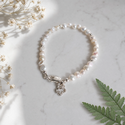 A beaded bracelet made of irregular white button pearls with a silver clasp and flower charm, styled on a white marble surface with dried flowers and a green fern leaf.