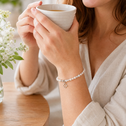 A woman in a cream-colored robe holds a white ceramic cup, showcasing a delicate white pearl bracelet with a small silver flower charm on her wrist.