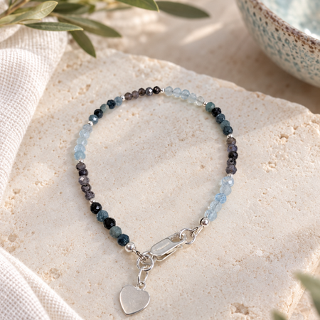 A delicate beaded bracelet with light blue, dark blue, and purple/gray beads, featuring a silver clasp and a small heart-shaped charm, displayed on a textured beige stone slab with a linen cloth and green leaves.
