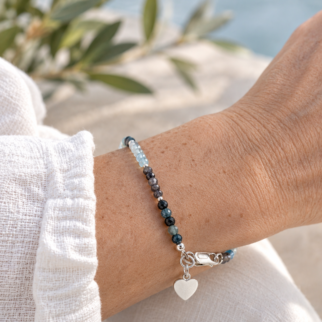A close-up of a person's wrist wearing a delicate beaded bracelet with dark blue and light blue beads, a silver clasp, and a white heart charm, styled with a white linen sleeve against a soft, blurred outdoor background.