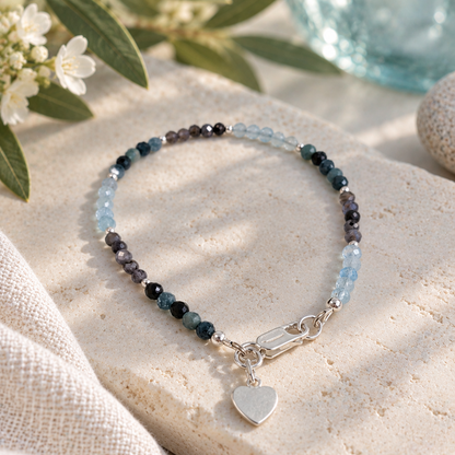 A beaded bracelet with a silver heart charm, displayed on a textured beige stone. The bracelet features faceted beads in shades of light blue, dark blue, and purple/gray, with a silver lobster clasp and a polished silver heart charm.