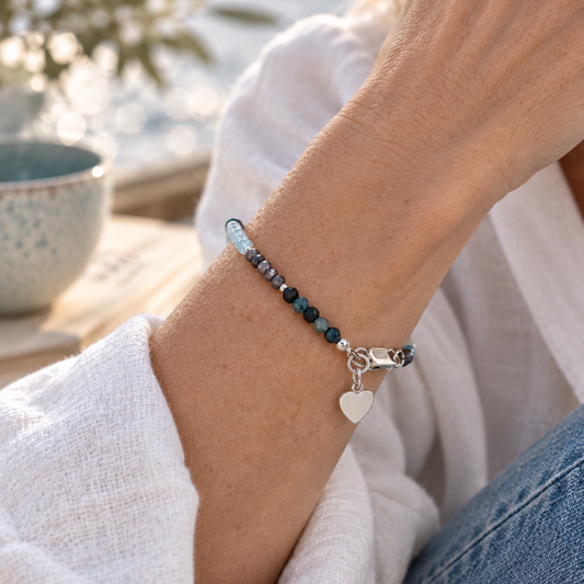 A close-up lifestyle shot of a person wearing a beaded bracelet with dark blue and silver beads, a silver lobster clasp, and a small silver heart charm, styled with a white linen shirt and blue jeans.