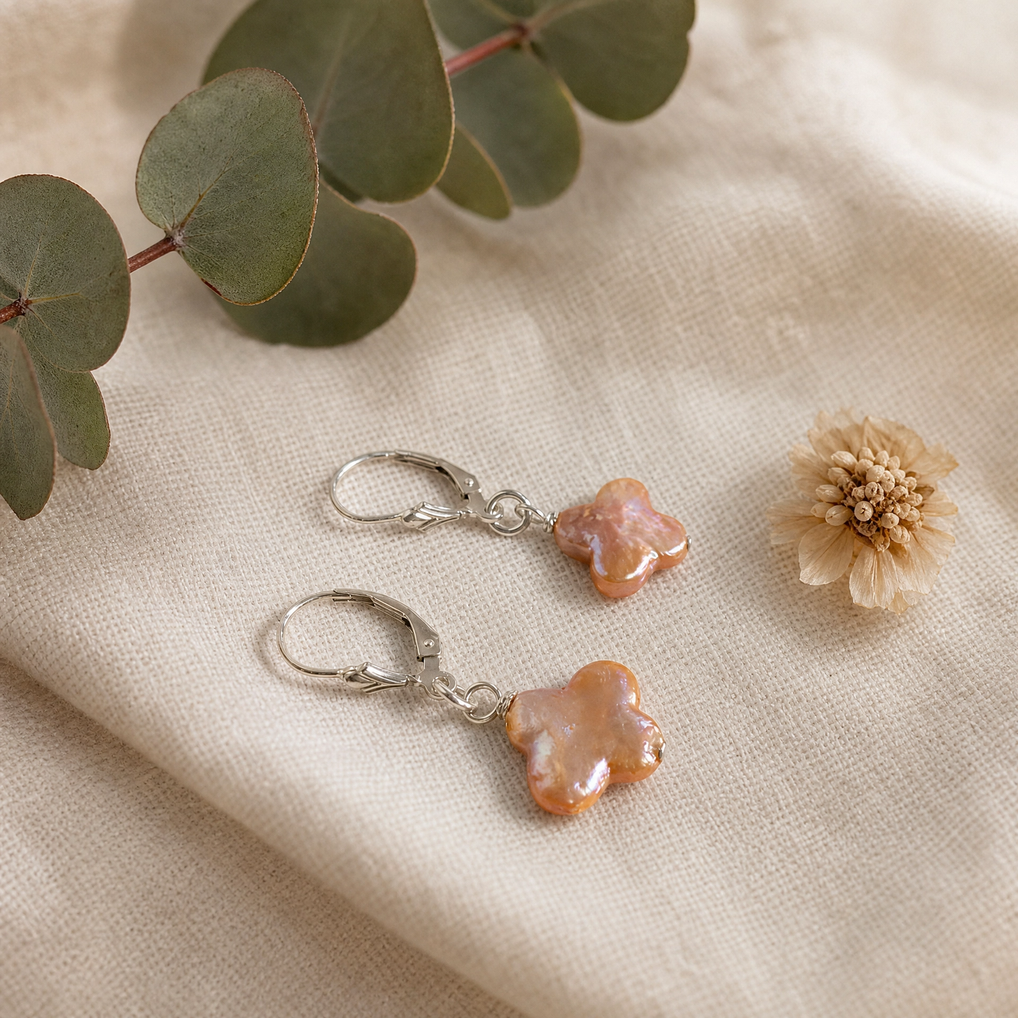 A pair of silver leverback earrings with irregular, glossy peach-orange clover pearls, styled on a beige linen fabric with eucalyptus leaves and a dried flower.