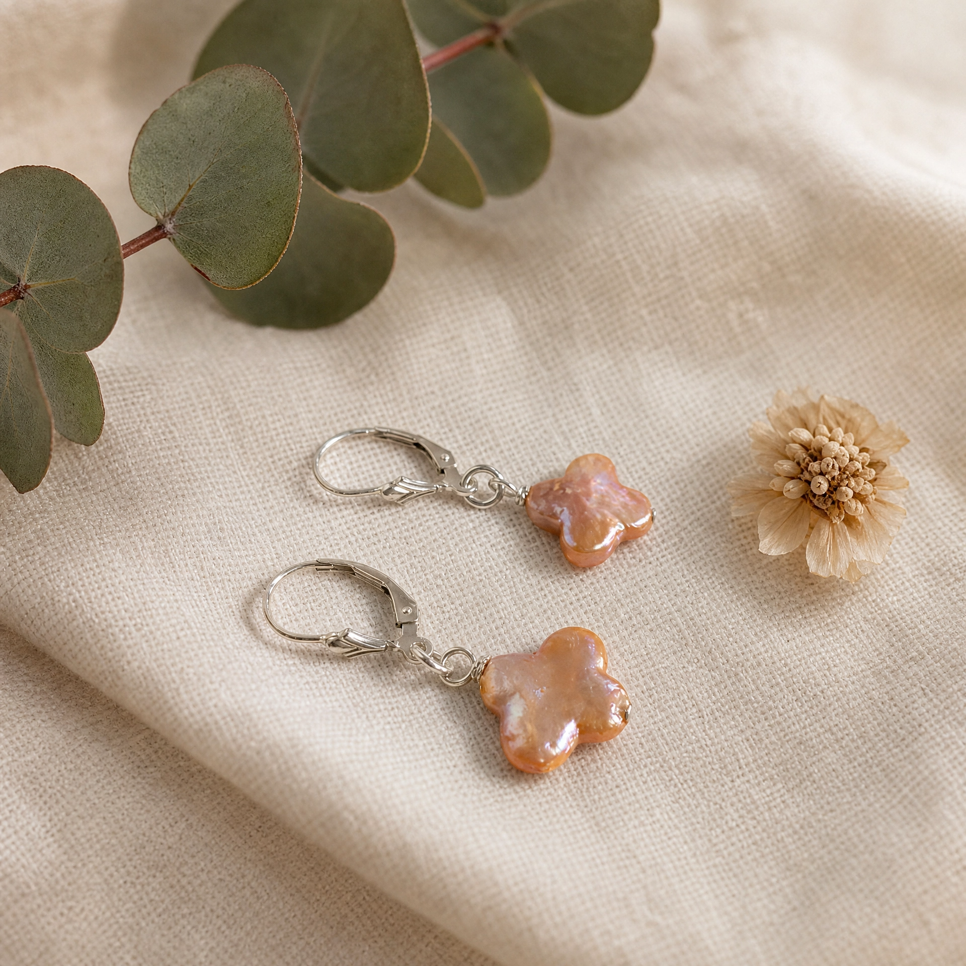 A pair of silver leverback earrings with irregular, glossy peach-orange clover pearls, styled on a beige linen fabric with eucalyptus leaves and a dried flower.